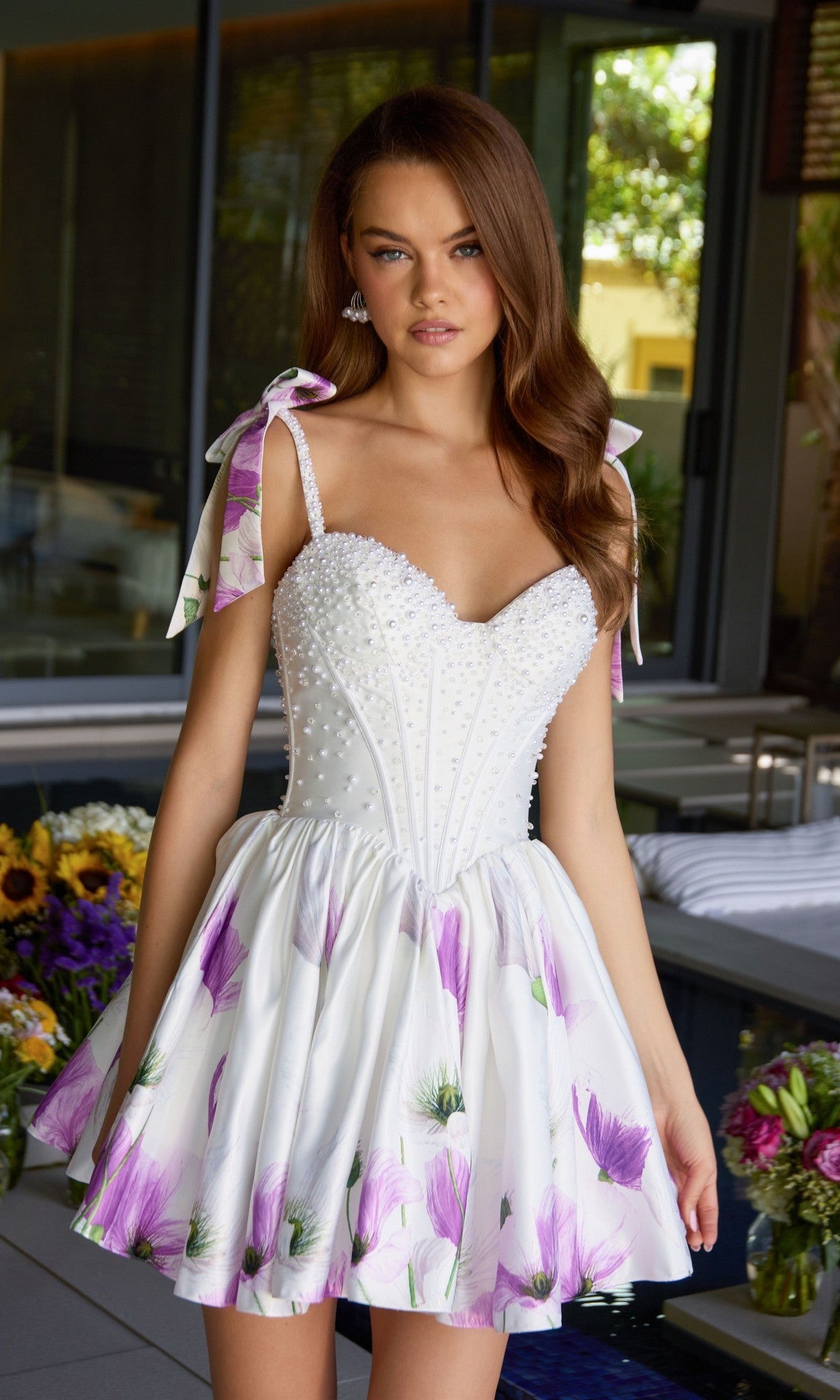 A woman stands indoors wearing the Ava Presley 42432 short cocktail dress, featuring a floral print satin fabric, pearl-embellished corset, and ribbon-tied shoulder straps. Flowers and greenery decorate the background.